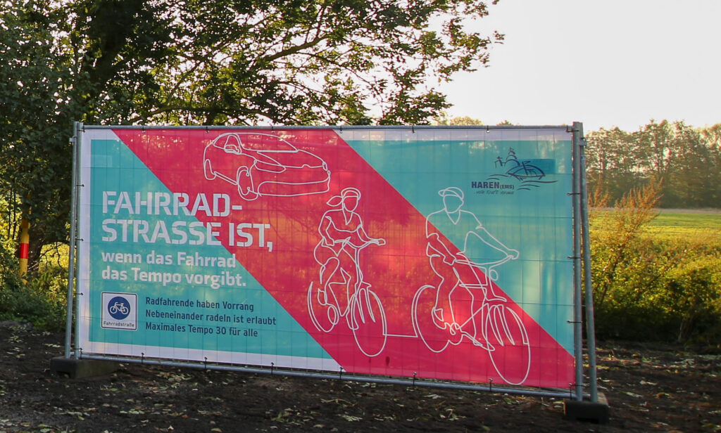 Zwei Bauzaunbanner entlang der Fahrradstraße weisen auf die wichtigsten Verkehrsregelungen einer Fahrradstraße hin. Vor allem, dass das Rad das Tempo vorgibt.