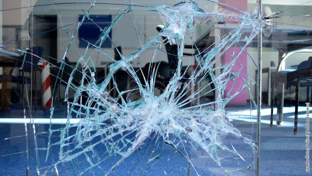 Zerbrochenes Glas mit starkem Einschlag in der Mitte, blauer Teppich im Hintergrund, Innenraum sichtbar.