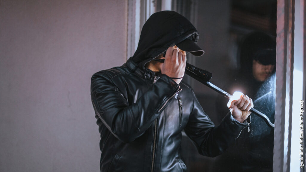 Einbrecher mit Brechstange vor Fenster im Dunkeln, trägt schwarze Kleidung, symbolisiert Sicherheit und Schutzmaßnahmen.