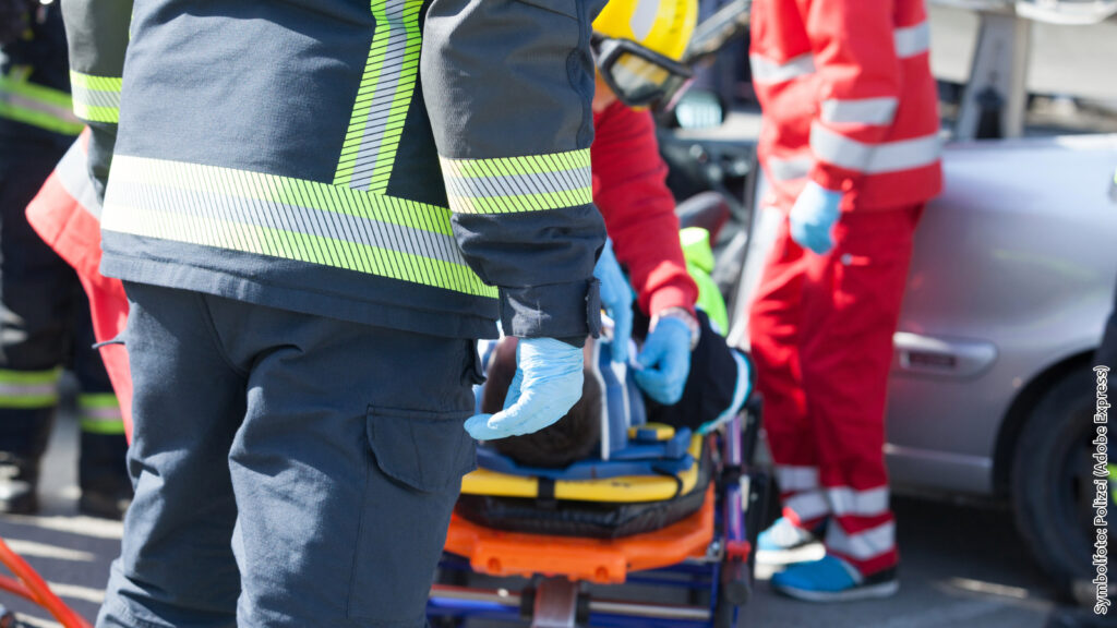 Rettungskräfte helfen verletzter Person auf Trage nach Autounfall. Polizei und Feuerwehr im Einsatz.