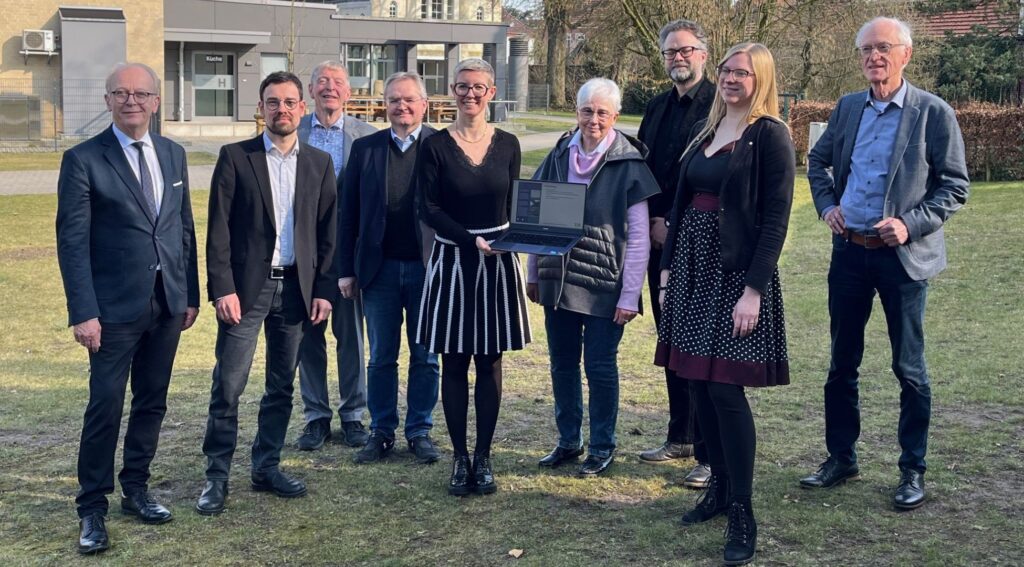 Gruppe von Menschen draußen, Frau in der Mitte hält Laptop, sonniger Tag, lächeln, Teamarbeit, Gebäude im Hintergrund.