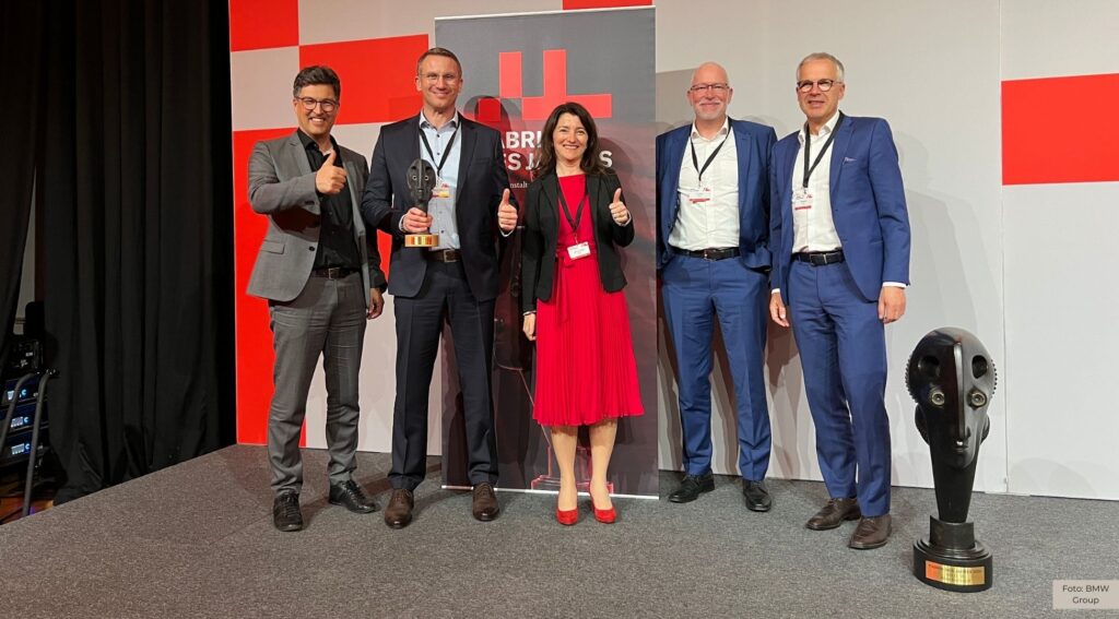 Gruppe von fünf Geschäftsleuten mit Auszeichnung auf Bühne. Zwei Männer zeigen Daumen hoch. Trophäe auf Boden.