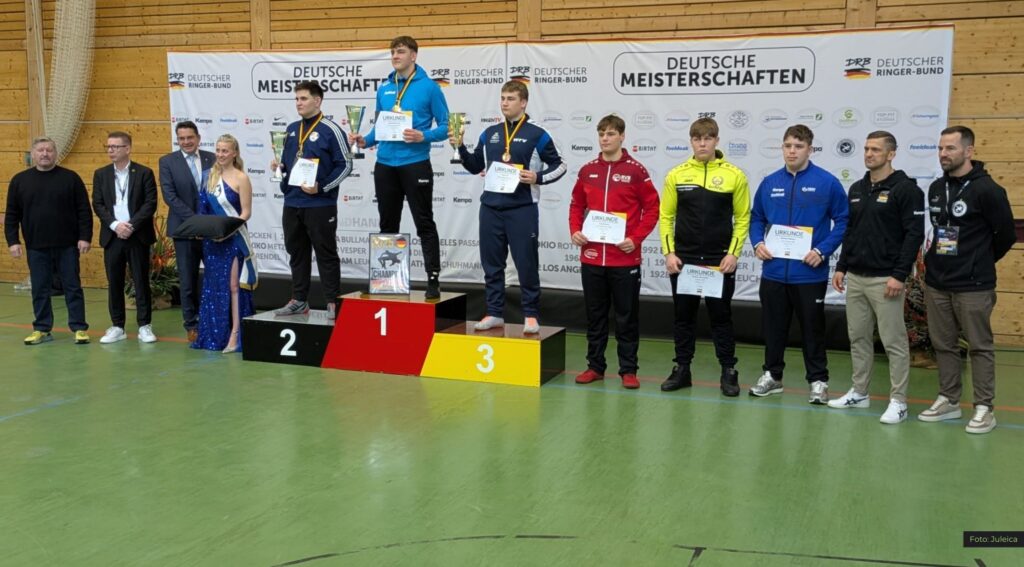 Siegerpodest bei deutscher Meisterschaft im Ringen, Sportler mit Urkunden und Pokalen, Gruppenfoto vor Banner.