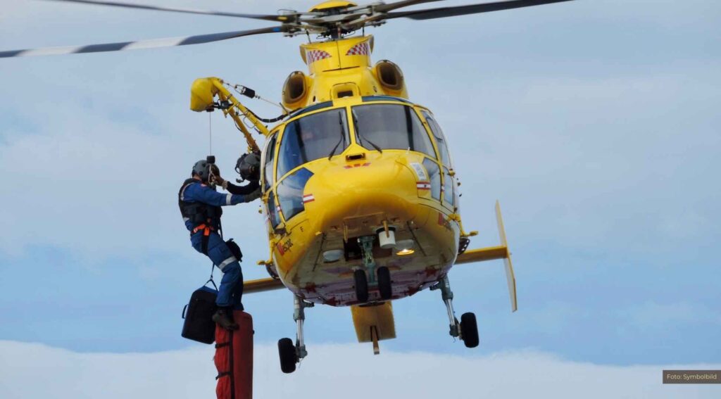 Gelber Rettungshubschrauber im Einsatz, während ein Retter mit Seilwinde heruntergelassen wird.