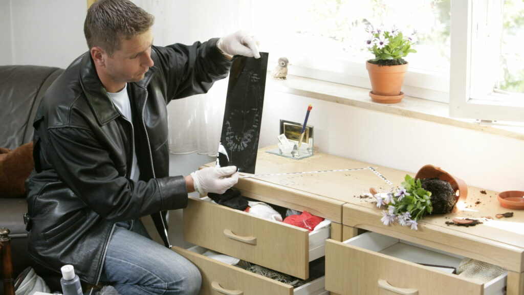 Mann untersucht Sicherheitsfolie in Wohnungsszene, offenbare Schubladen und zerbrochener Blumentopf im Vordergrund.
