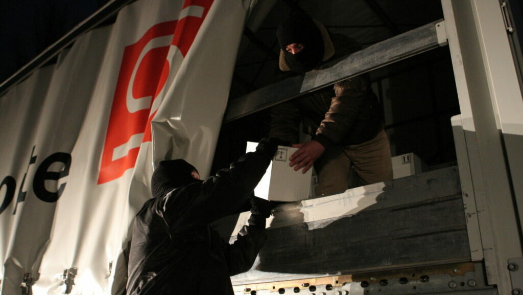 Zwei Personen in dunkler Kleidung laden bei Nacht Kartons von einem LKW aus.