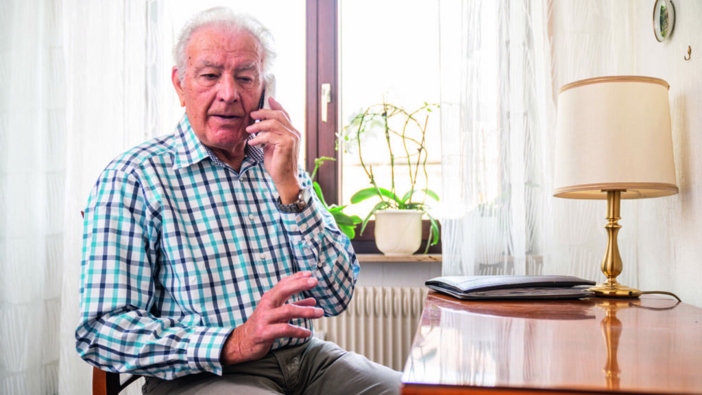 Älterer Mann telefoniert in hellem Raum, neben ihm eine Lampe und Pflanzen auf dem Tisch.