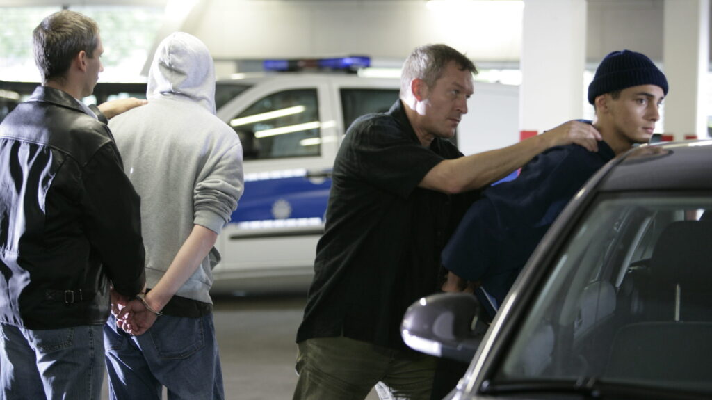 Polizeiverhaftung: Zwei Männer werden von Zivilpolizisten an einem Fahrzeug festgenommen. Einsatzfahrzeug im Hintergrund.