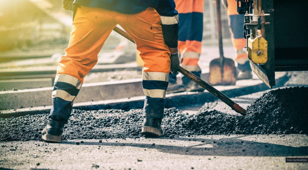 Straßenbauarbeiter verdichten frischen Asphalt bei Sonnenschein.
