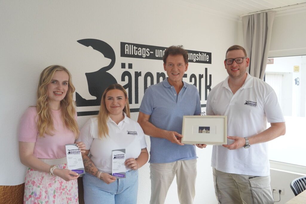 Gruppe von vier Personen mit Bärenstark-Logo an der Wand, zwei halten Flyer, einer ein Bild in einem Büro.