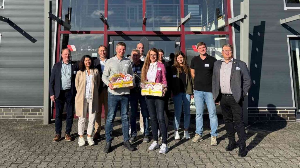 Gruppe von Menschen vor einem Gebäude, zwei halten Präsentkörbe, lächelnd in die Kamera bei sonnigem Wetter.