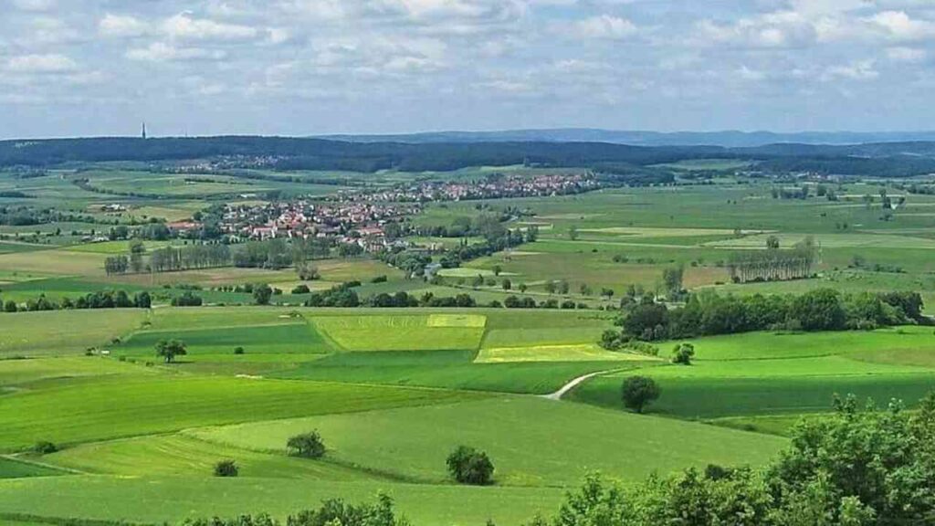 Landschaftspanorama mit grünen Feldern, Hügeln und einem Dorf im Hintergrund unter blauem Himmel.