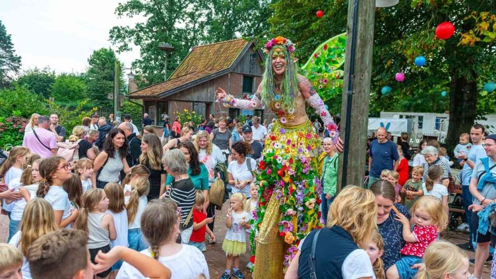 Bunte Straßenparade mit bunt verkleideter Person auf Stelzen, umgeben von lächelnden Kindern und Erwachsenen in festlicher Stimmung.
