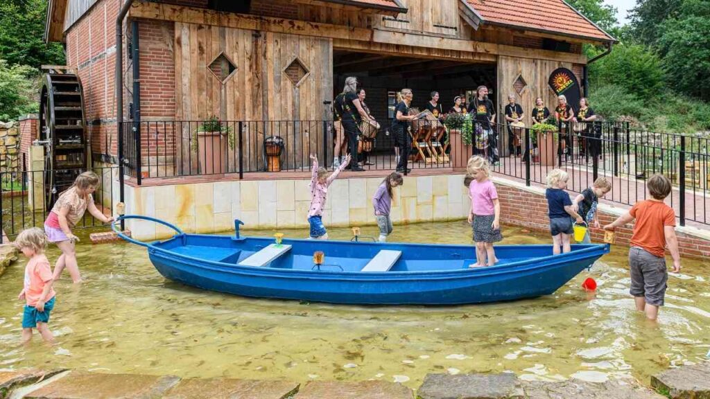 Kinder spielen im Wasser vor einer Holzmühle, während eine Musikgruppe im Hintergrund trommelt. Fröhliche Sommeratmosphäre.
