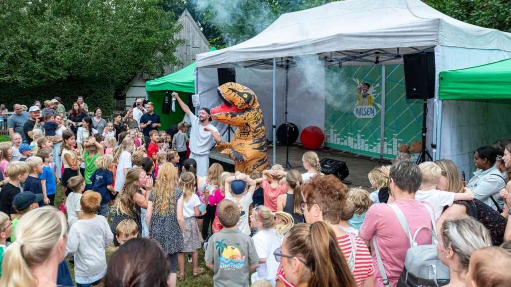 Kinderveranstaltung mit Dinosaurier-Auftritt im Freien, viele Kinder und Erwachsene schauen begeistert auf das Spektakel.