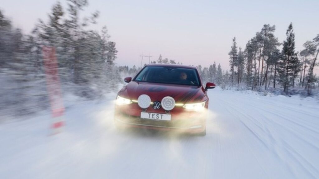 Rotes Auto fährt auf verschneiter Straße durch winterlichen Wald, klarer Himmel im Hintergrund.