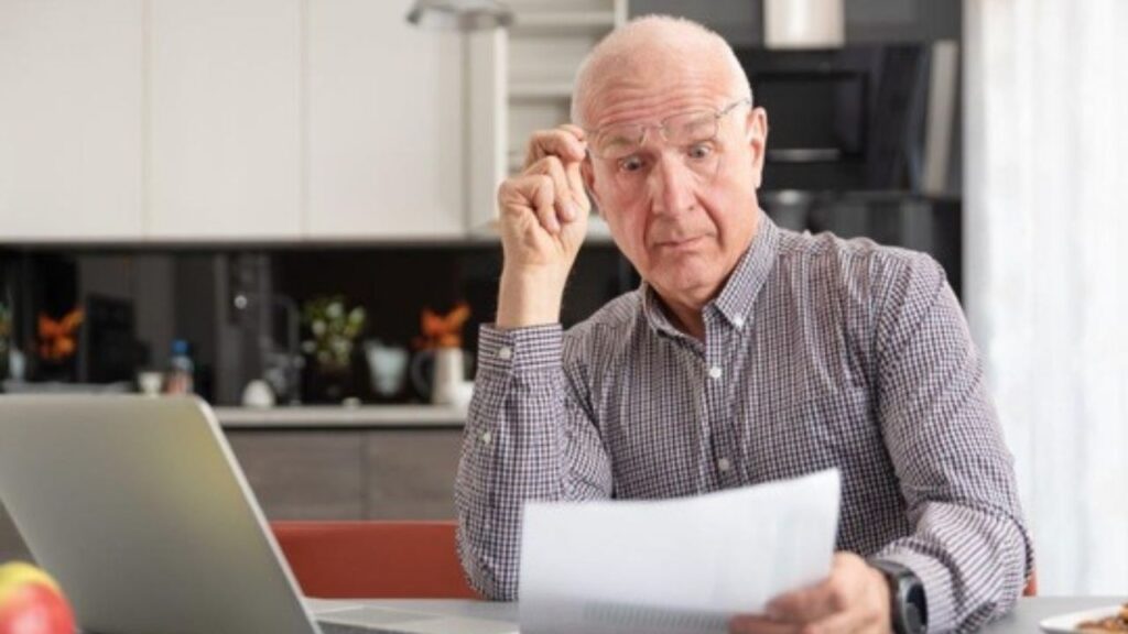 Älterer Mann liest Dokument, sitzt vor Laptop in moderner Küche, nachdenklich, mit Brille in der Hand.