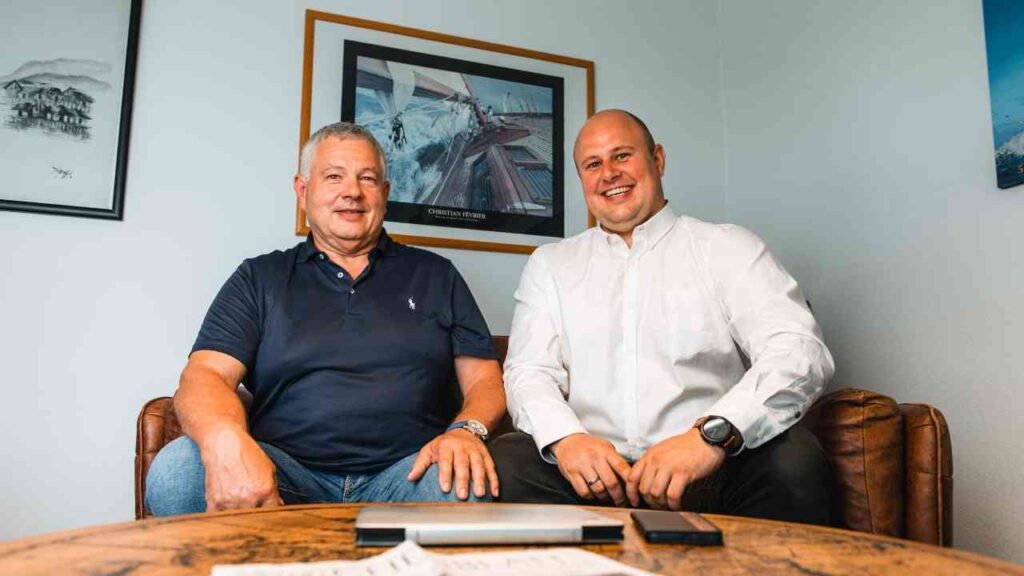 Zwei lächelnde Männer sitzen auf Bürostühlen, hinter ihnen Kunstwerke, im Gespräch über Business-Themen.