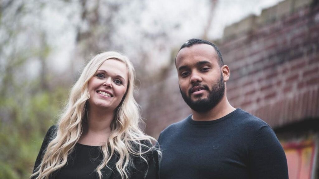 Ich weiß nicht, wer auf dem Bild ist, aber hier ist ein passender Alt-Text: Lächelndes Paar in Freizeitkleidung vor Backsteinmauer.