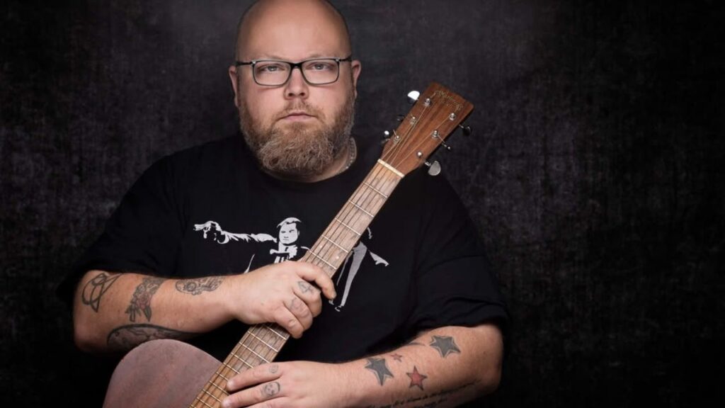 Andreas Kümmert mit Brille und Tattoos hält Akustikgitarre vor dunklem Hintergrund.