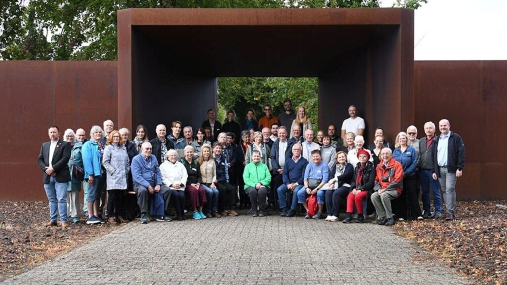 Gruppe von Menschen vor modernem, rechteckigem Denkmal im Freien.