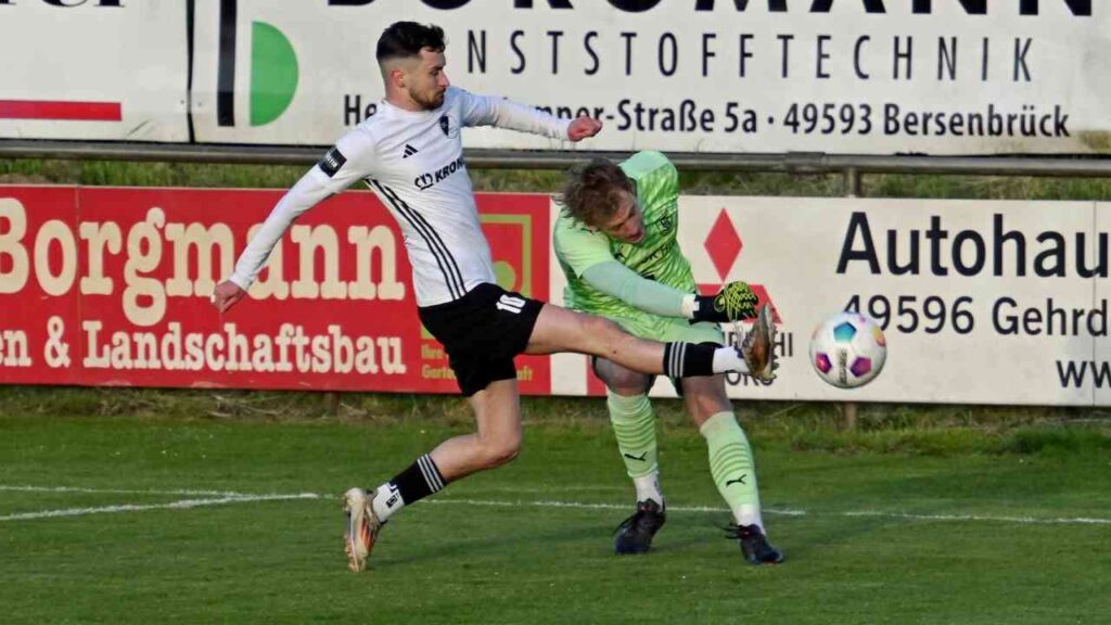 Ein Fußballspieler im weißen Trikot schießt den Ball, während der Torwart versucht, ihn zu blocken.
