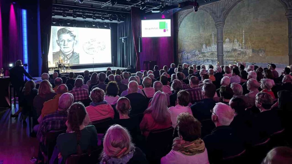 Publikum bei einer Präsentation in einem Theater, Leinwand zeigt historisches Bild, lebhafte Atmosphäre, voller Saal.