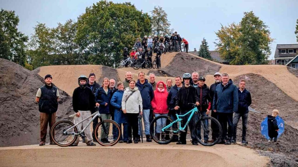 Gruppe von Menschen posiert mit Fahrrädern vor einer BMX-Strecke, umgeben von naturbelassenen Erdhügeln und Bäumen.