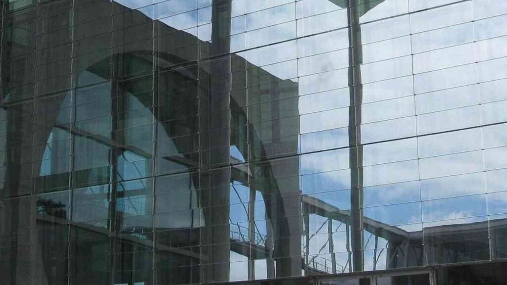 Moderne Glasfassade mit spiegelnder Architektur im Hintergrund unter blauem Himmel.