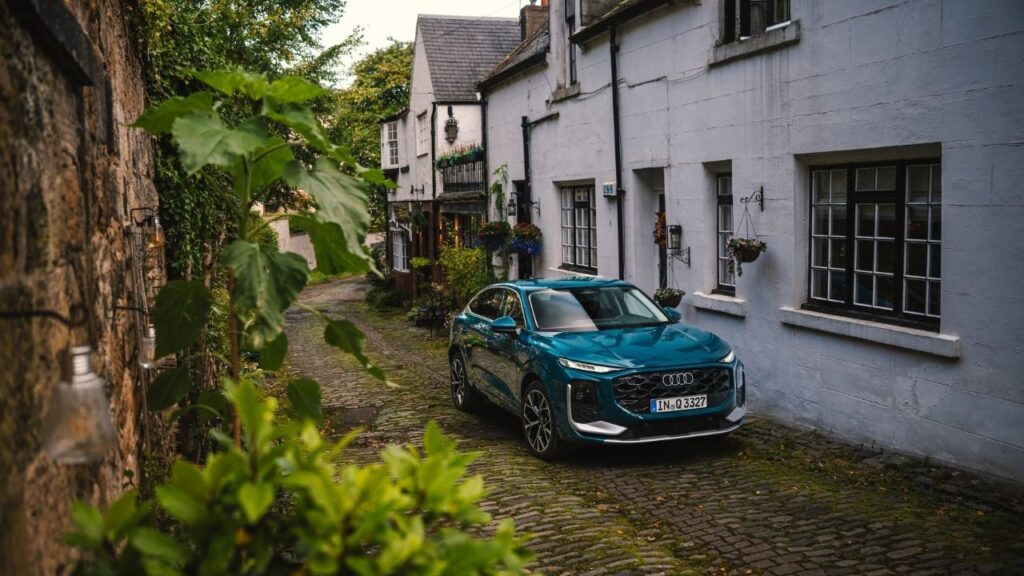 Blauer Audi SUV in enger, gepflasterter Altstadtgasse mit historischen Gebäuden und üppigem Grün.