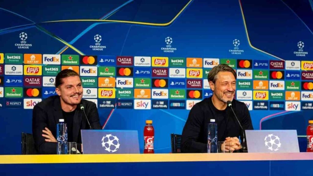 Zwei Männer bei einer Champions League Pressekonferenz, lächeln vor Mikrofonen und Sponsorenwand.