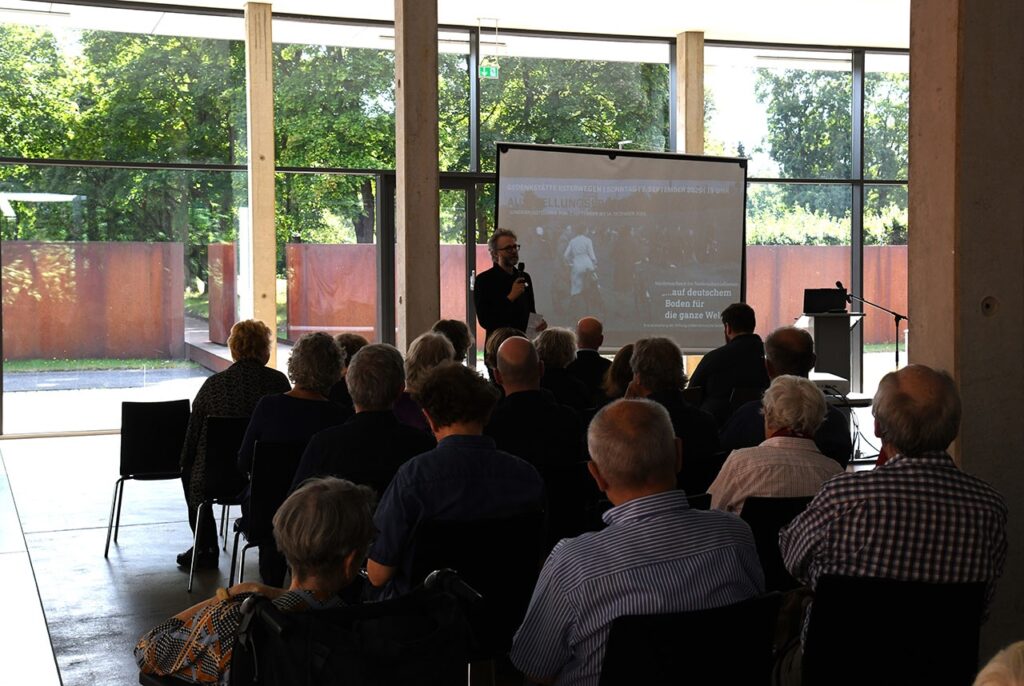 Person hält Vortrag vor Publikum in modernem Gebäude, Projektion im Hintergrund, Gedenkstätte Estewergen.