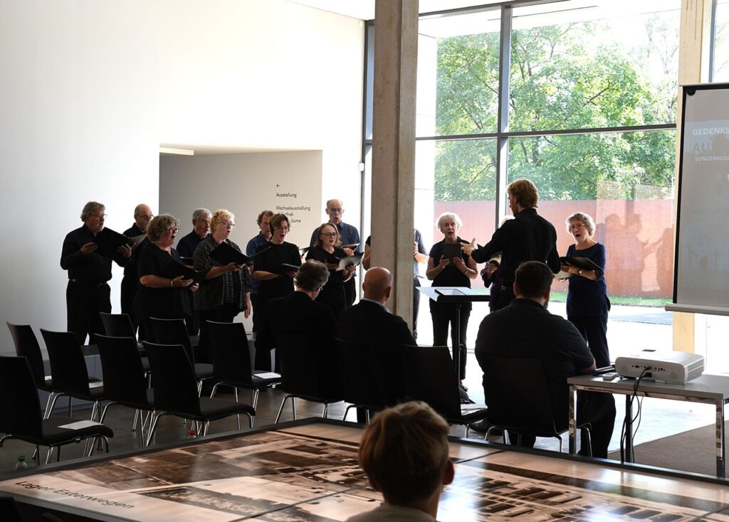 Chor singt in moderner, lichtdurchfluteter Ausstellungshalle, dirigiert von einer Person. Publikum hört aufmerksam zu.