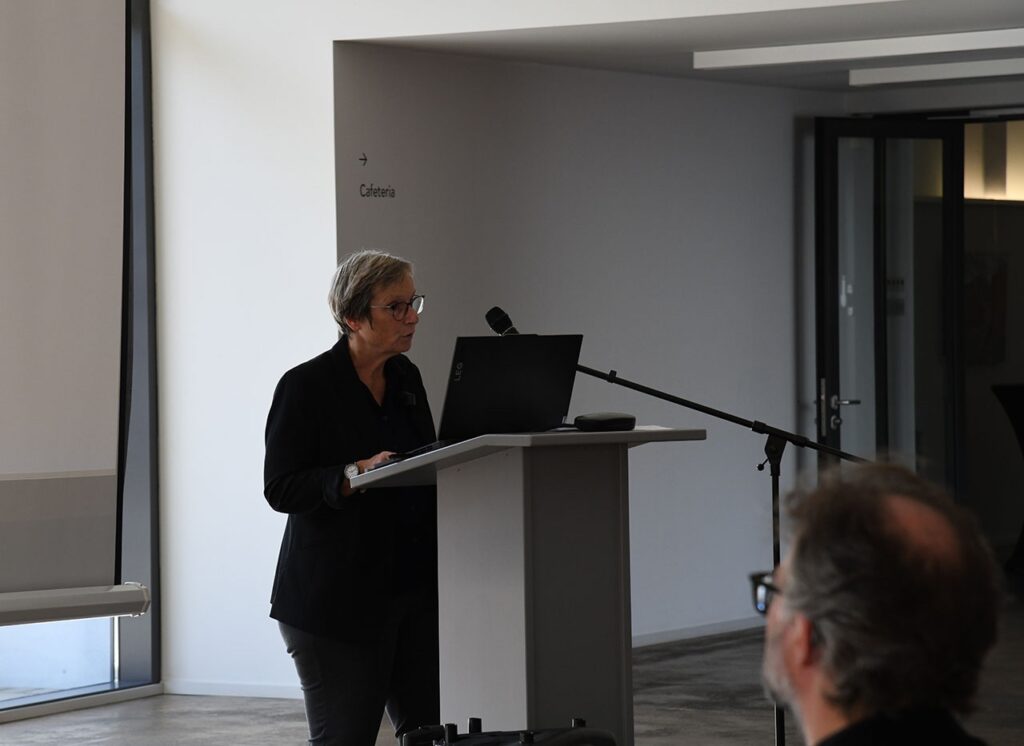 Frau hält Vortrag am Rednerpult mit Laptop und Mikrofon, im modernen Konferenzraum. Zeichen zur Cafeteria im Hintergrund.