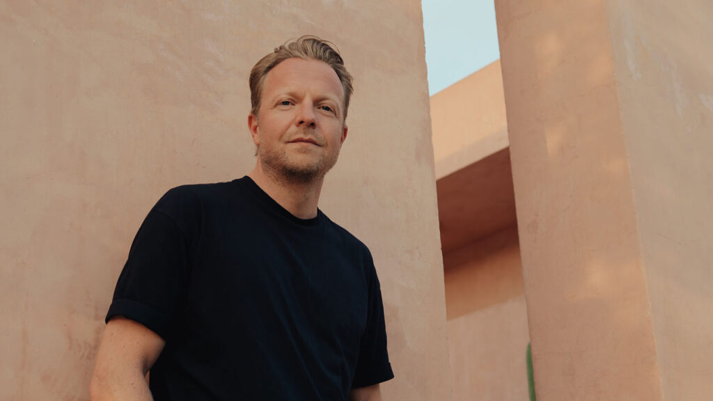 Christoph Dassler in schwarzem T-Shirt lehnt lässig an einer beigen Wand bei sonnigem Wetter, modernes Architekturdesign im Hintergrund.