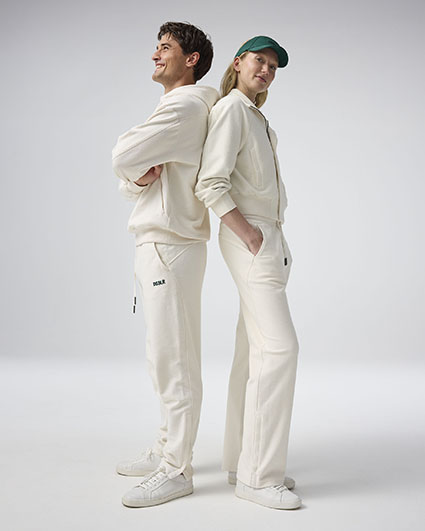 Zwei Personen in weißer Sportbekleidung stehen Rücken an Rücken auf hellem Hintergrund. Beide tragen weiße Sneaker.