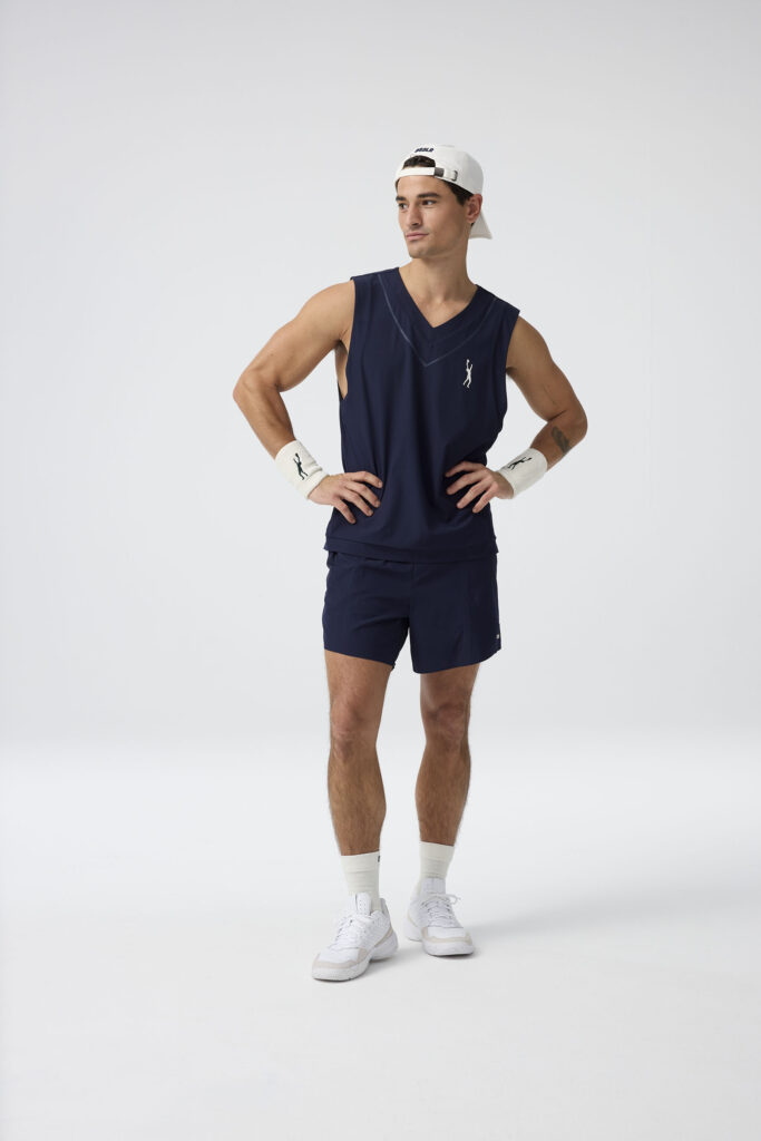 Mann in Sportkleidung posiert selbstbewusst vor weißem Hintergrund. Sportoutfit: Dunkles Trikot, Shorts und Baseballkappe.