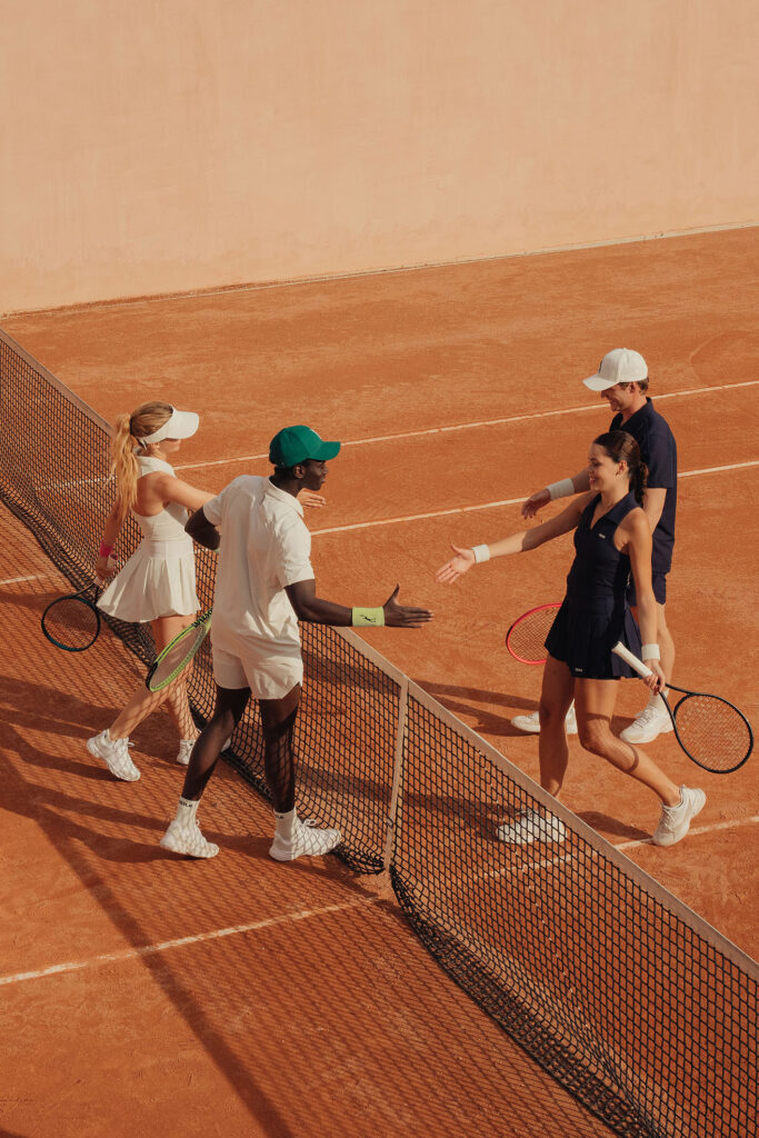 Tennisspieler reichen sich nach dem Spiel die Hand auf einem Sandplatz. Zwei Männer, zwei Frauen, strahlendes Wetter.
