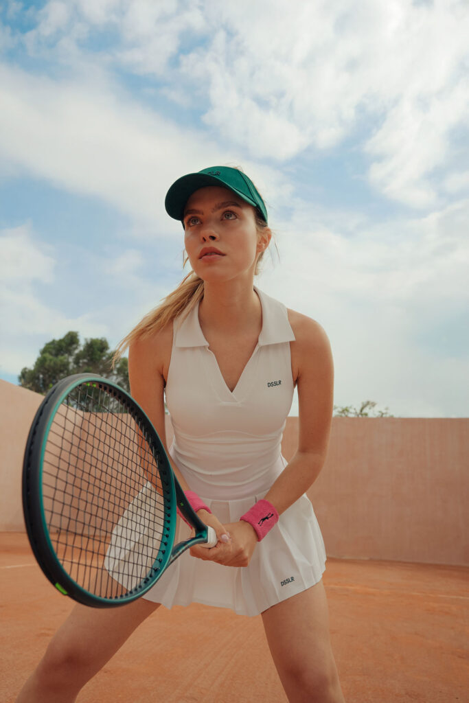 Frau in weißem Tennisoutfit und grünem Visor, bereit für den Aufschlag auf Sandplatz unter blauem Himmel.