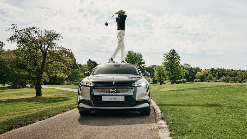 Person golft vom Dach eines Autos auf einem malerischen Golfplatz mit Bäumen und Wolken am Himmel im Hintergrund.