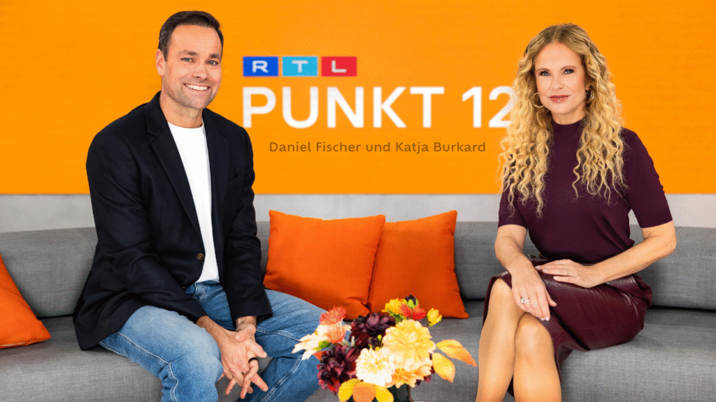 Moderatoren vor RTL Punkt 12-Logo auf grauem Sofa, lächeln in Kamera, orangefarbener Hintergrund.