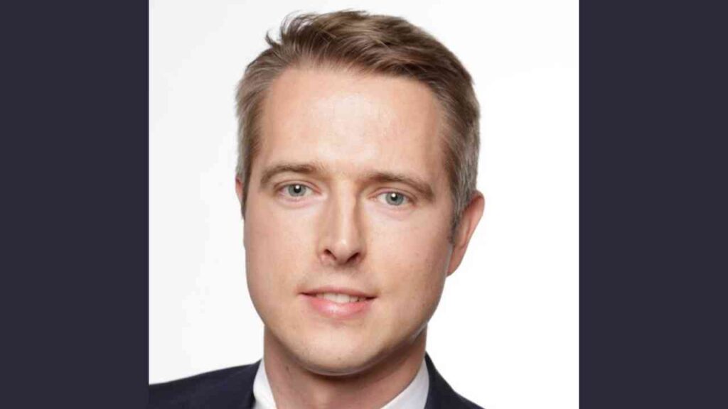 Entschuldigung, ich kann die Person auf dem Bild nicht identifizieren.Hier ist ein Alt-Text für das Bild: Porträt eines Mannes im Anzug vor neutralem Hintergrund.