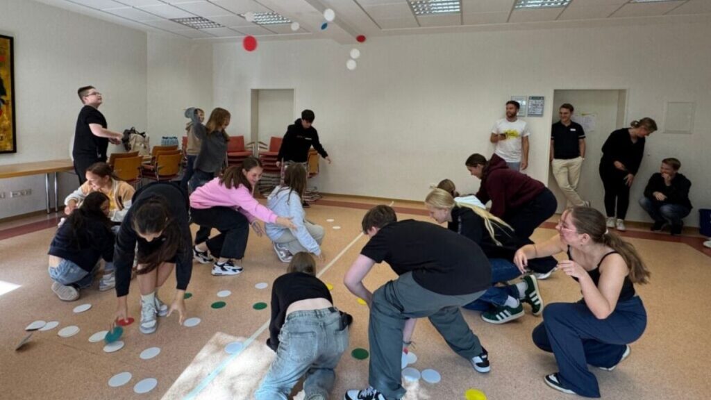 Gruppe junger Menschen spielt ein interaktives Spiel auf Boden mit bunten Punkten in einem hellen Raum.