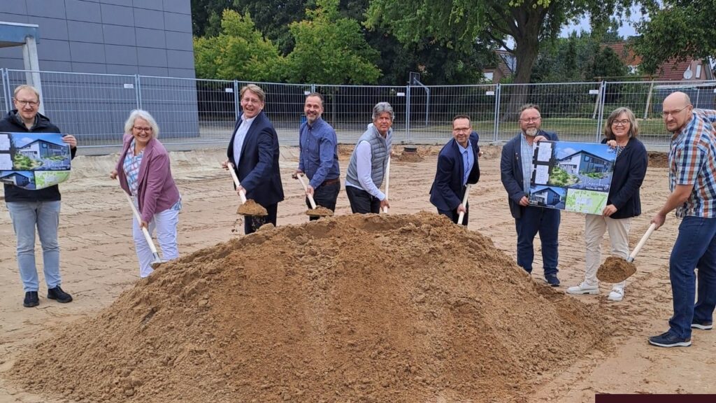 Menschen beim Spatenstich für ein Bauprojekt, lächeln in die Kamera, Bauplakate im Hintergrund.