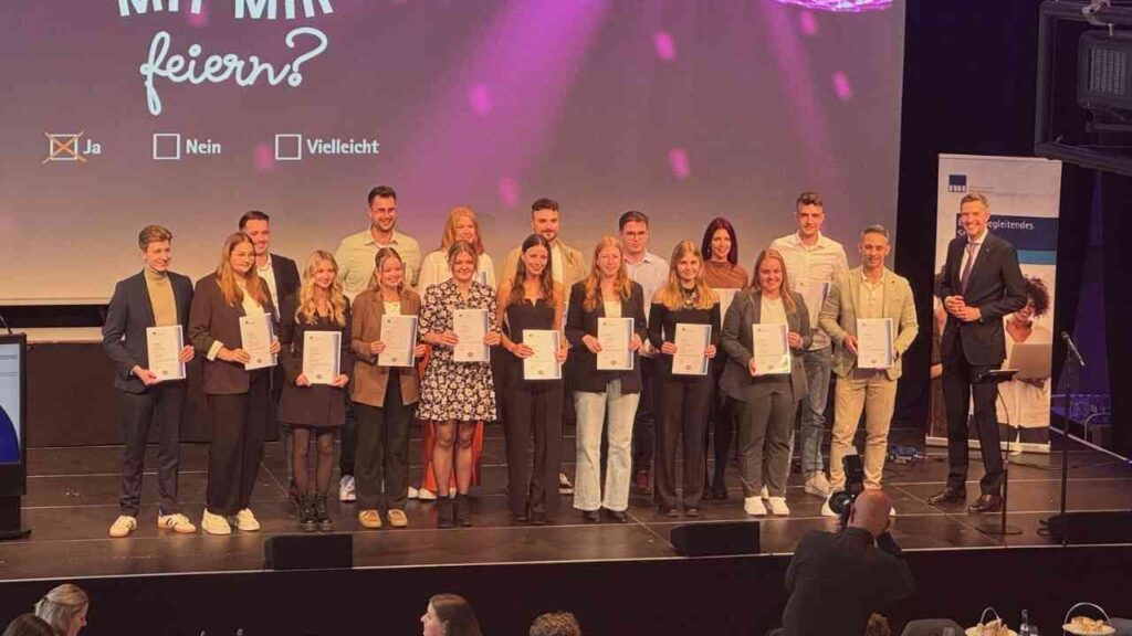 Eine Gruppe von Menschen steht auf einer Bühne und hält Zertifikate bei einer Abschlussveranstaltung.