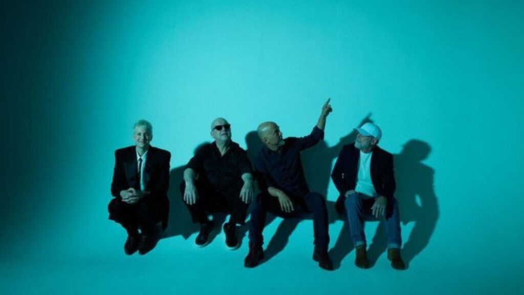 Vier Männer sitzen vor einer blauen Wand, einer zeigt nach oben.