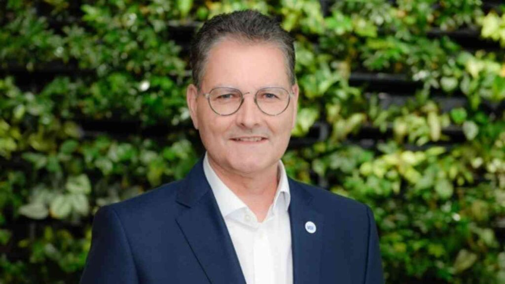 Porträt eines Mannes im Anzug vor einer grünen Pflanzenwand, lächelnd und mit Brille.