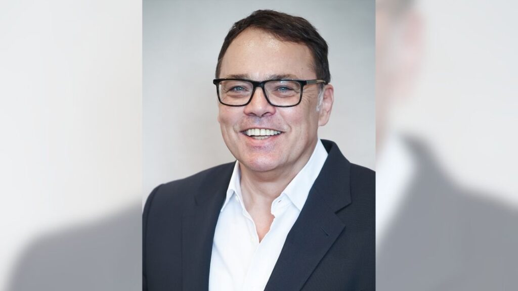 Lächelnder Mann mit Brille in Anzug, vor neutralem Hintergrund. Geschäftsprofil oder professionelles Porträtfoto.