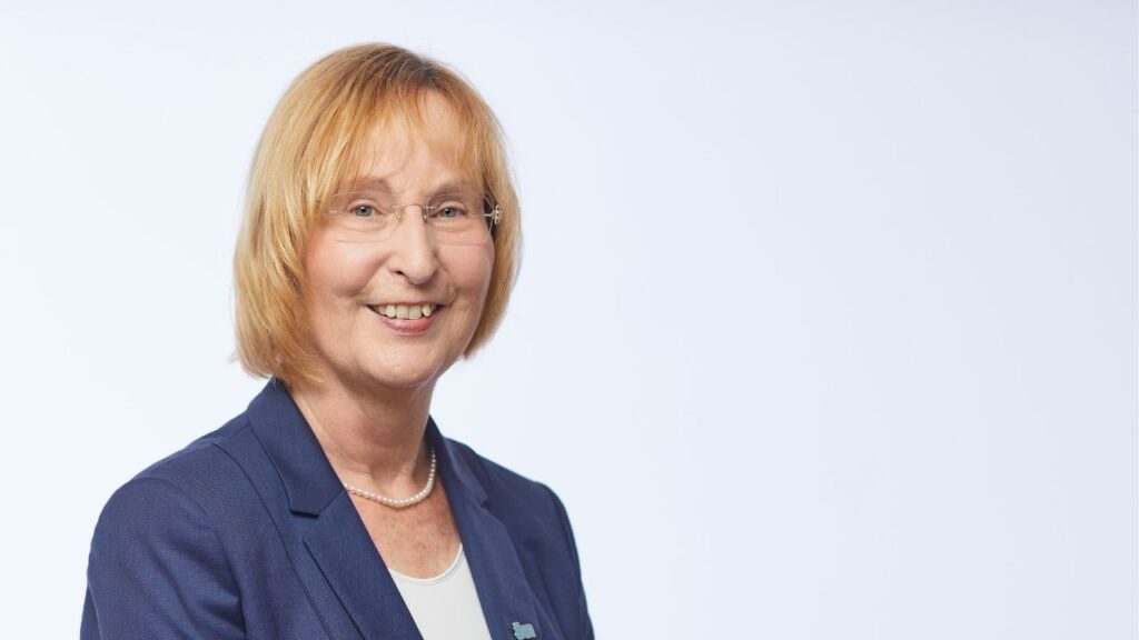 Porträtfoto einer lächelnden Frau mit blondem Haar und Brille, gekleidet in einem dunkelblauen Blazer vor einem hellen Hintergrund.