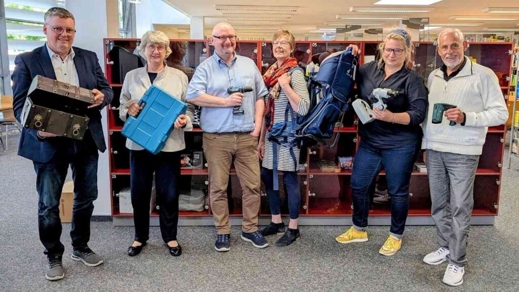 Gruppe von sechs Menschen mit Werkzeug in einer Bibliothek, lächelnd vor Schließfächern stehend.